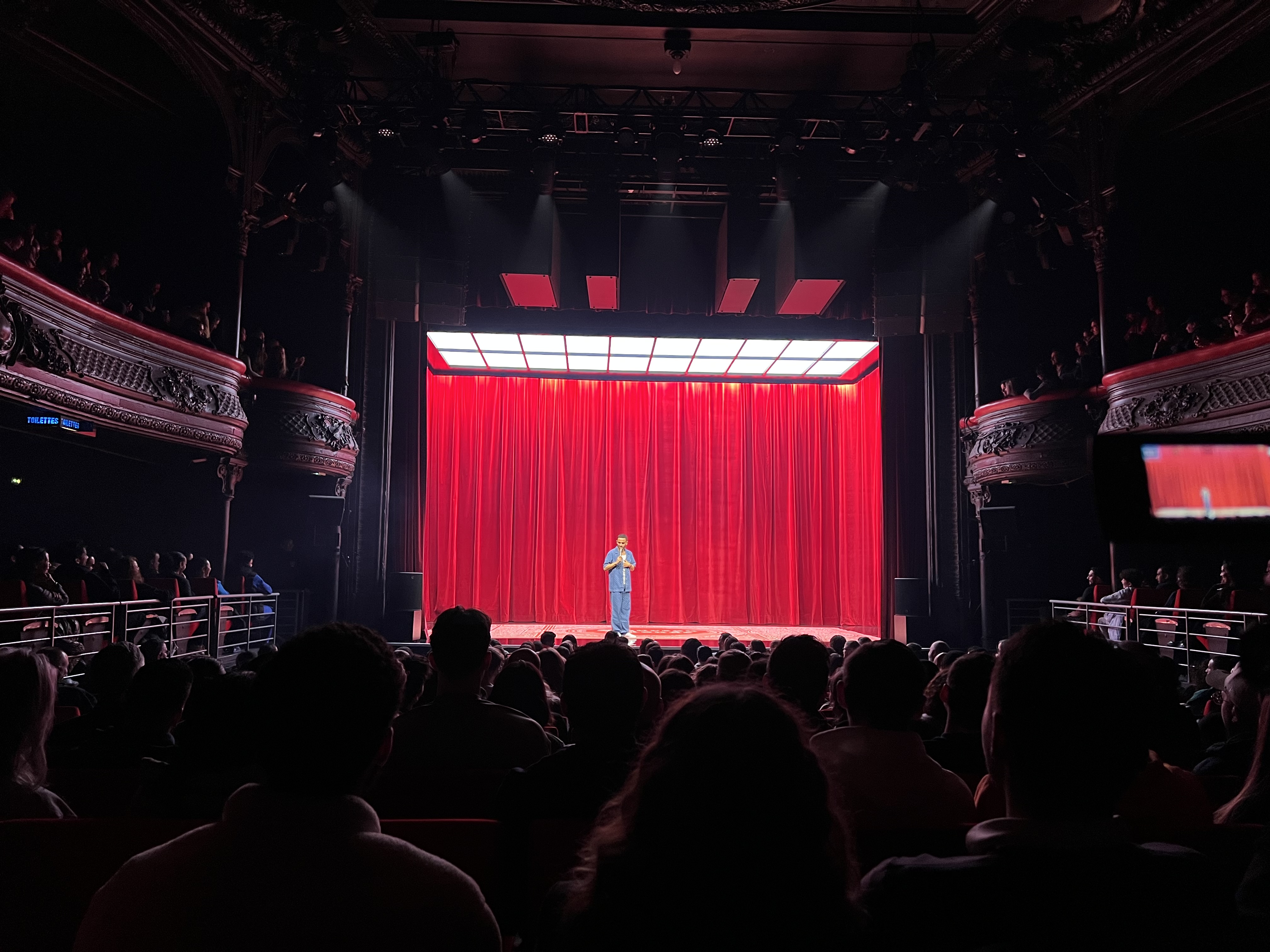 Malik Bentalha sur scène devant le rideau rouge et le plafond lumineux de Nouveau Monde