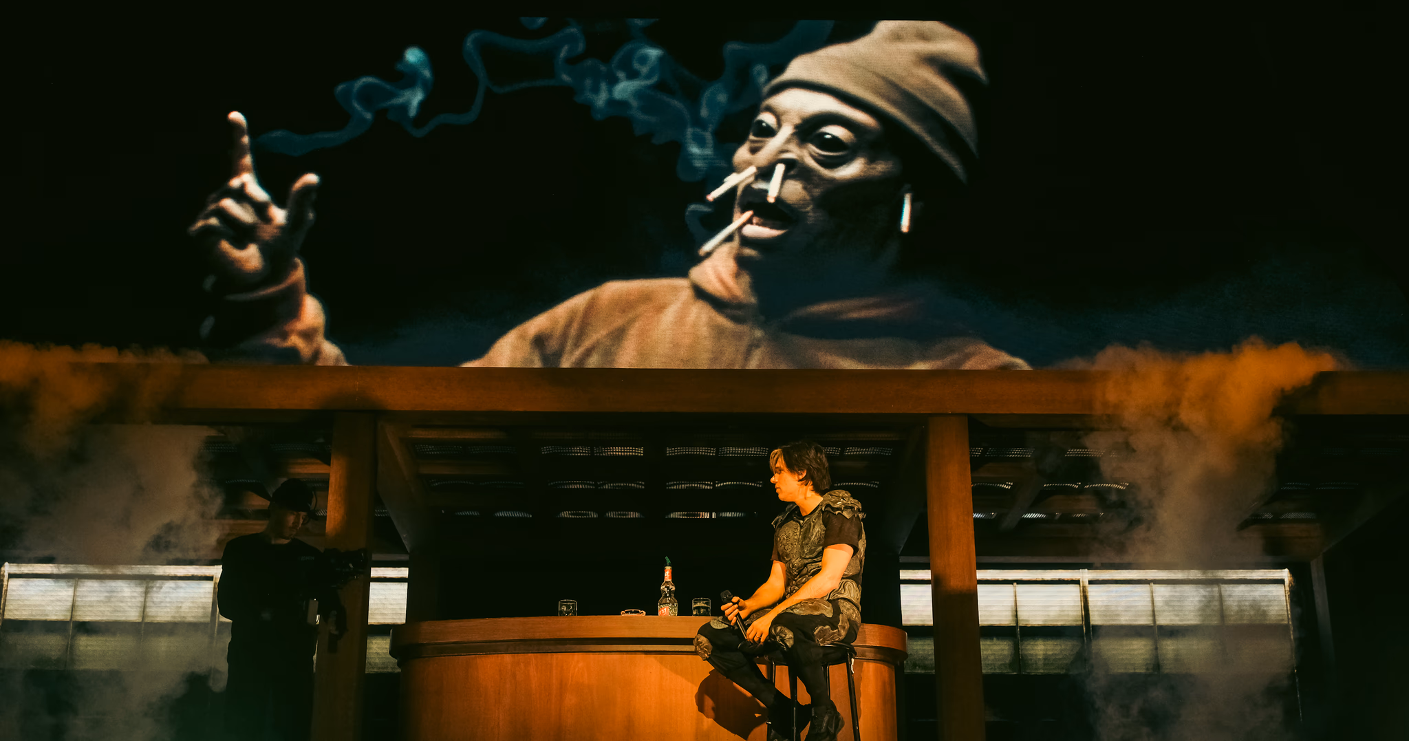 Orelsan assis au bar du décor sous une projection vidéo